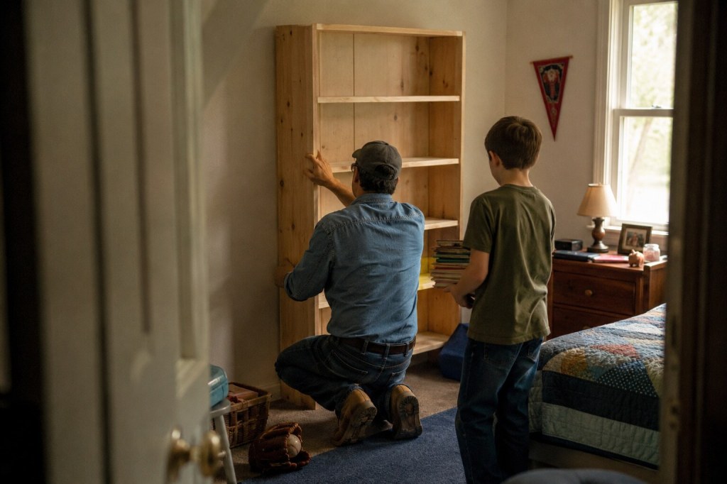 A father installs a wooden bookshelf in his son's bedroom while the boy waits with a stack of books.