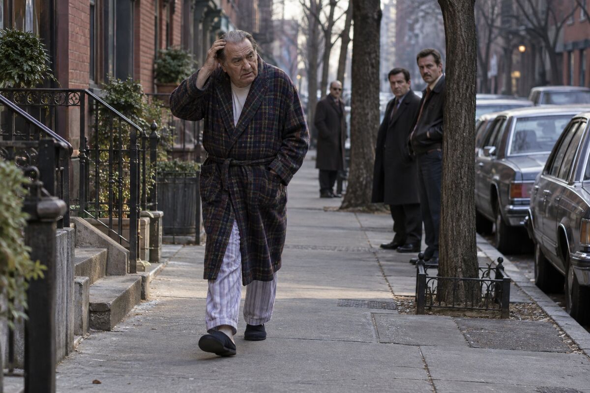 An older man in a bathrobe and pajamas walking through Greenwich Village while observers watch from a distance.