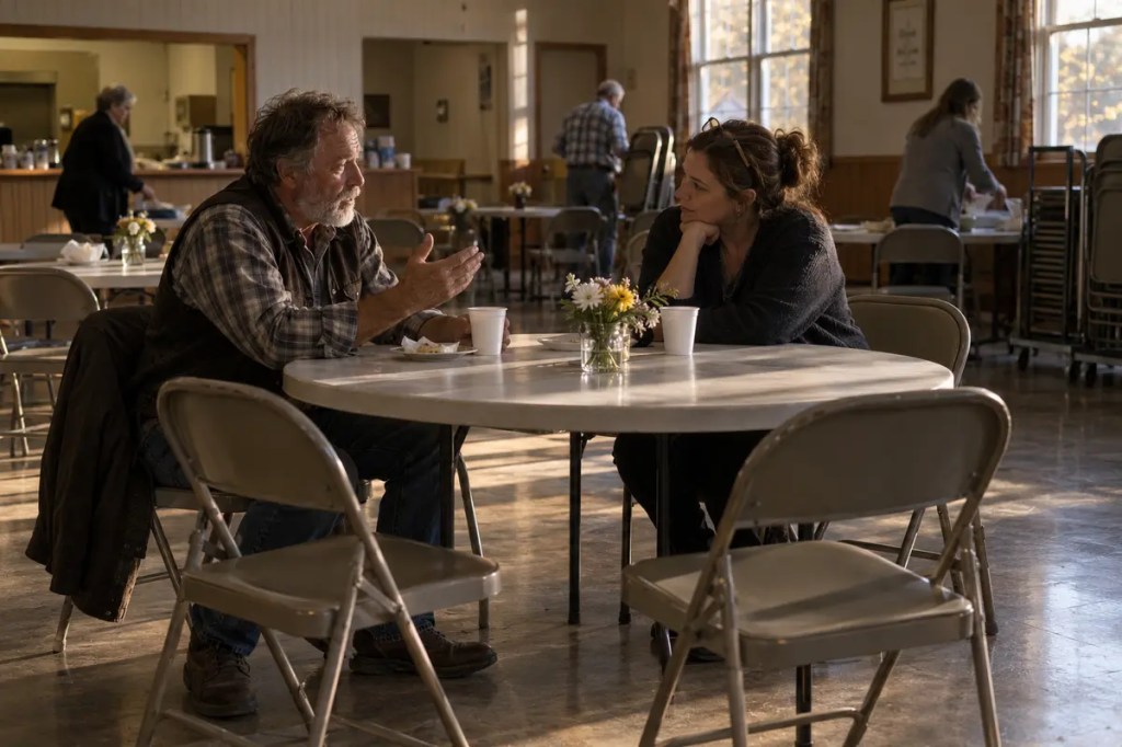 Two people remain in a quiet church fellowship hall after a memorial gathering, sharing a story across a table.